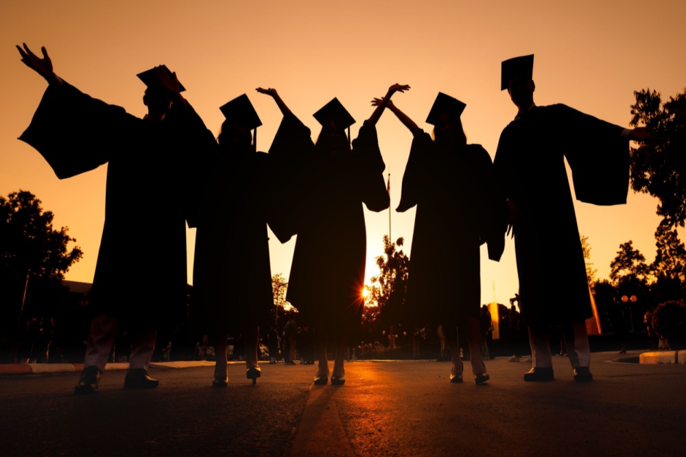 Dual enrollment: Black silhouette of 5 young adult in graduation gowns and caps standing with arms upraised against completely orange sunset sky