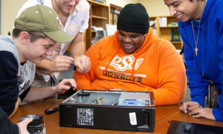 High school students cyber-defense: A young man in an orange sweatshirt sits behind an open computer case. Two young people sit on either side of him.