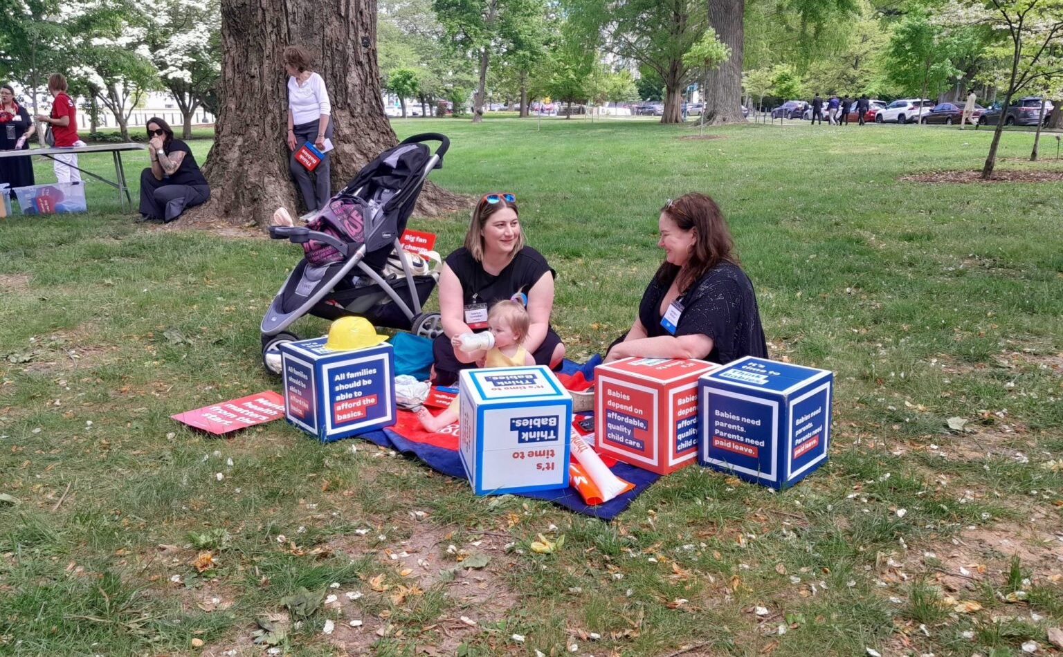 Parents tote toddlers to D.C. for child tax credit and child care: two women, one with a baby, sitting and talking on a blanket in a park with protest materials