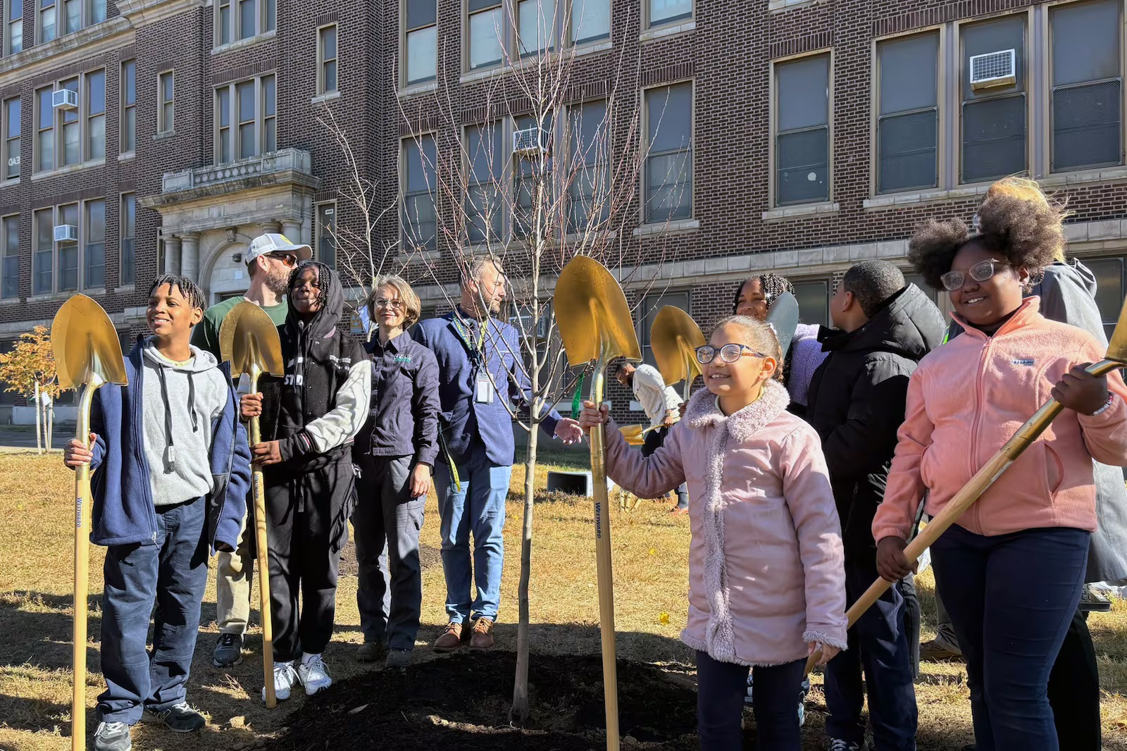 Philadelphia schools get state grant to plant more trees: young students with shovels stand and smile in front of newly planted tree outside school building