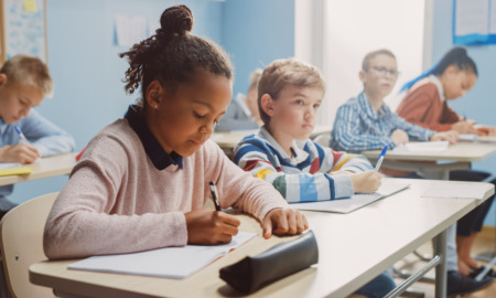 Pandemic School Funding: # elementary students cit in a row ar desks writing in papers while listening to teacher