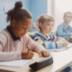 Pandemic School Funding: # elementary students cit in a row ar desks writing in papers while listening to teacher