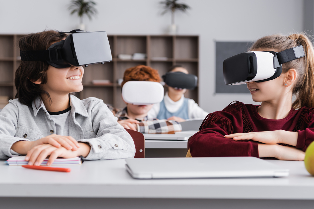 EdTech report: Happy elementary classmates looking at each other while sitting at desks gaming in vr headsets in classroom