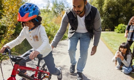 Parenting in America today report: dad helping daughter on bike learn to ride while other daughter films on phone off to the side and mom watches on