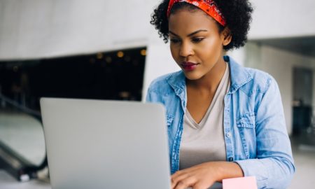 Focused young African female working online with a laptop while sitting at a table in the lobby of a modern office building