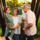 Kinship foster care: One older man and one older woman both with gray hair stand outside with grennery in front of green trees, the woman holding a dark-haired, female todder in sage green jumpsuit on her hip as girls waves at camera.