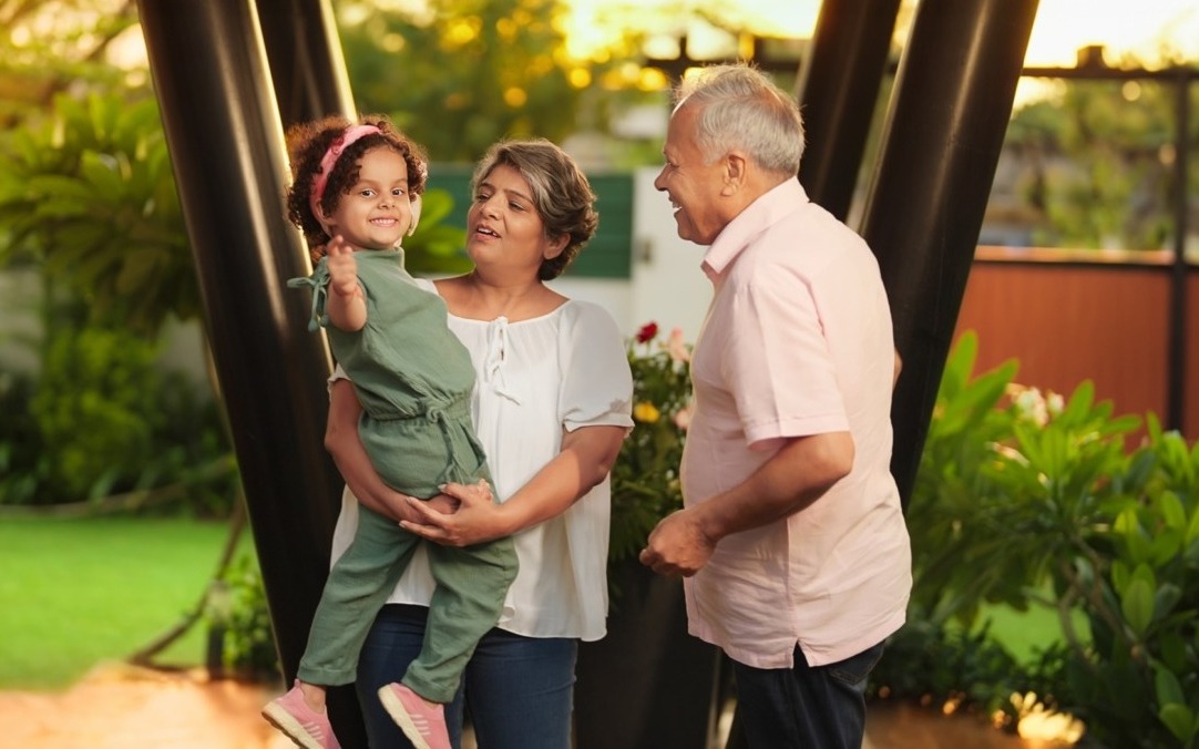 Kinship foster care: One older man and one older woman both with gray hair stand outside with grennery in front of green trees, the woman holding a dark-haired, female todder in sage green jumpsuit on her hip as girls waves at camera.