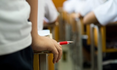 Schools face pressure to take harder line on discipline: closeup of students hand holding red pen with class in desks in background