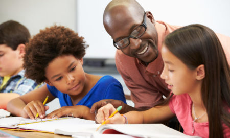 addressing student absenteeism, tutoring and afterschool programs: three pre-teen students sit working in notebooks while bald black man with dark glasses and red shirt explains lesson