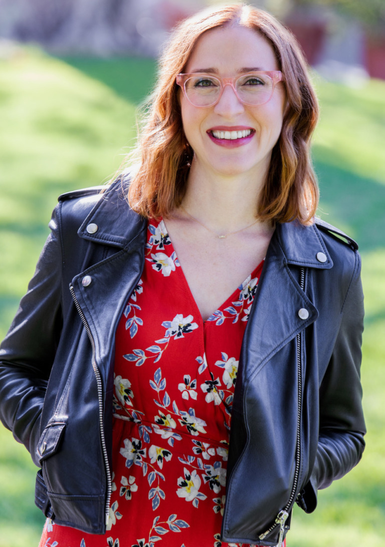 Rebecca Goldberg headshot: Wiman with long light brown hair in denin jacket and red dress stands in green field with hands in pockets