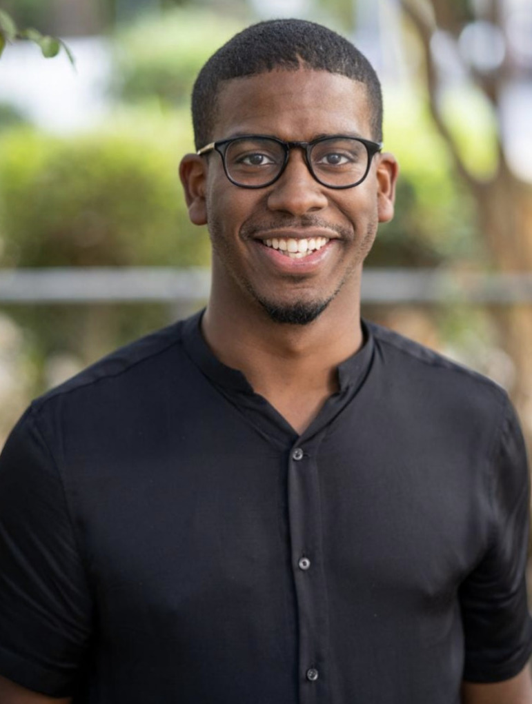 Sixto Cancel Headshot: Black man with shrt black hair and dark hoenrim glasses uv black shirt smiles into camera