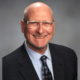 Afterschool California: Michael Funk Headshot bald older man with wirerim glasses in navy suit, light blue shirt and navy tie