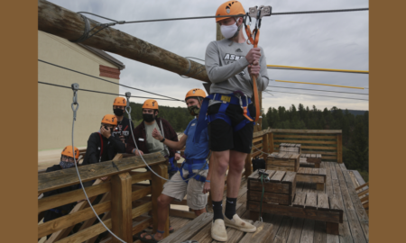 summer camp pandemic precautions: Young man Kole Linville in dark shorts, light shirt and yellow hardhat stands ready to take off on zipline wth other young adults lined up behind him