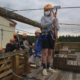 summer camp pandemic precautions: Young man Kole Linville in dark shorts, light shirt and yellow hardhat stands ready to take off on zipline wth other young adults lined up behind him