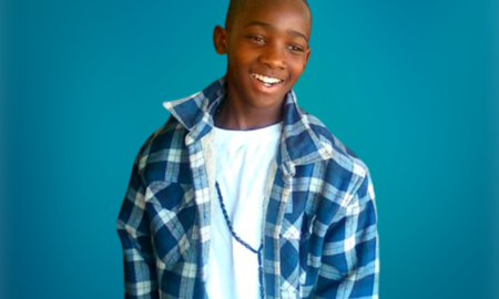 LaKeith Re-Sentencing: Young, black male teen with very short black hair in blue and white plaid long-sleeved shirt over white t-shirt stands in front of turquoise wall smiling broadly into the camera.