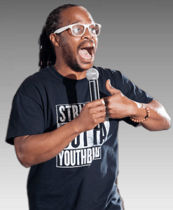 Jamiel Alexander Headshot: Black manwith long hair and  clear-framed glasses in black t-shirtstands speaking with hands acoss chest as fists meetingt